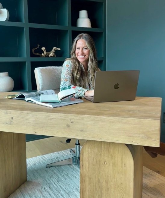 Natalie Mayes seated at her desk in front of a wall of deep teal built-in shelves